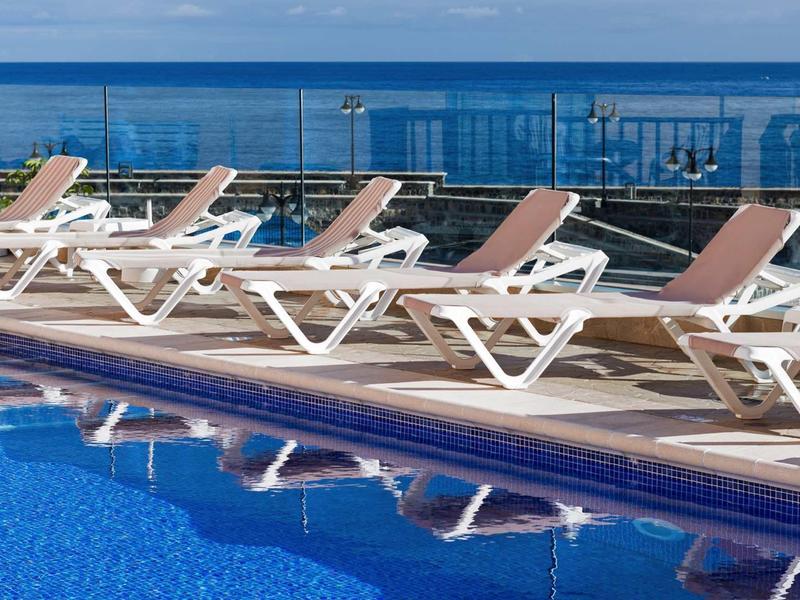 Reihe weißer Liegestühle mit braunen Polstern entlang eines blauen Pools am Meer.