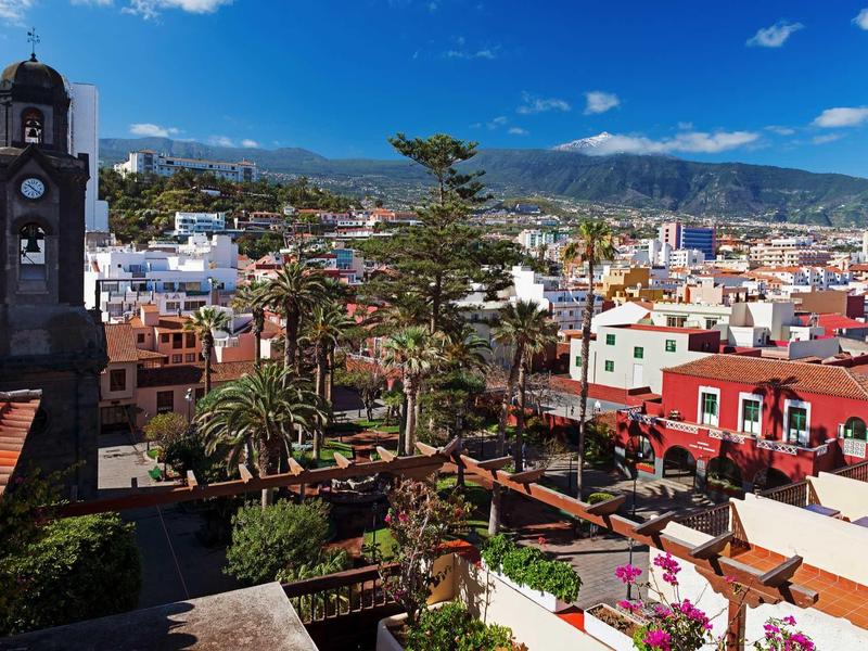 Stadt mit bunter Architektur, grünen Bäumen und Bergen im Hintergrund unter blauem Himmel.