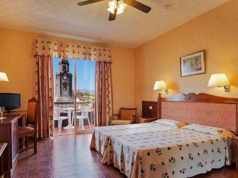 Helles Schlafzimmer mit Doppelbett, Holzmöbeln, Ventilator, Tisch, Stühlen und Balkon mit Blick.