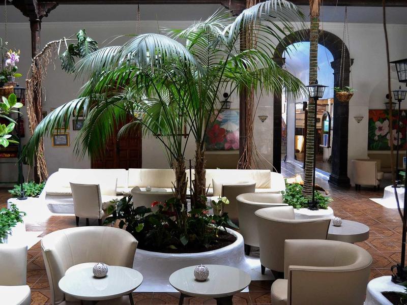Innenhof mit Palmen, weißen Sesseln und kleinen runden Tischen, umgeben von hohen Wänden und Bögen.