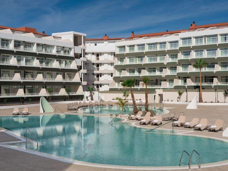 Nowoczesny kompleks hotelowy z basenem na zewnątrz, leżakami i niebieskim niebem w tle.