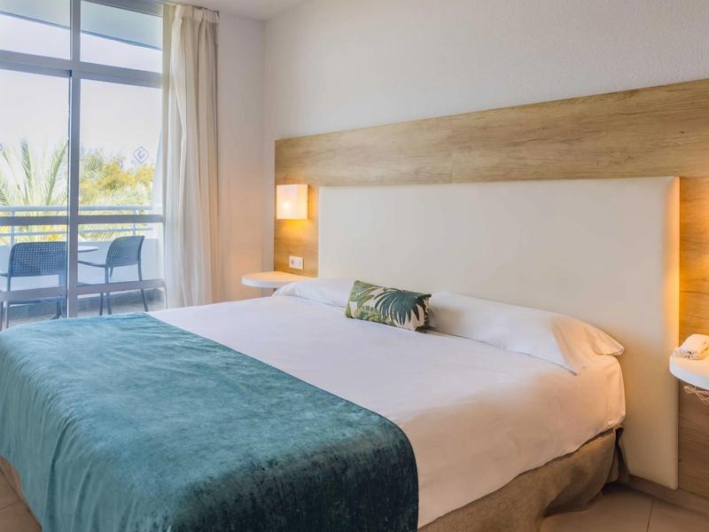 Jasny pokój hotelowy z dużym łóżkiem, drewnianym wystrojem i balkonem z krzesłami.