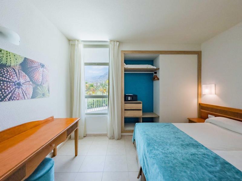 Jasny, nowoczesny pokój hotelowy z łóżkiem podwójnym, drewnianymi meblami i widokiem z okna.