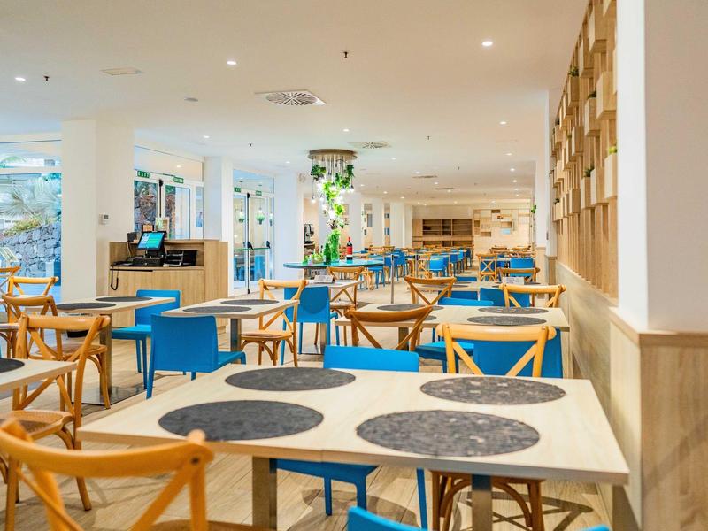 Jasna restauracja hotelowa z drewnianymi meblami, białymi stołami i niebieskimi krzesłami, światło dzienne.