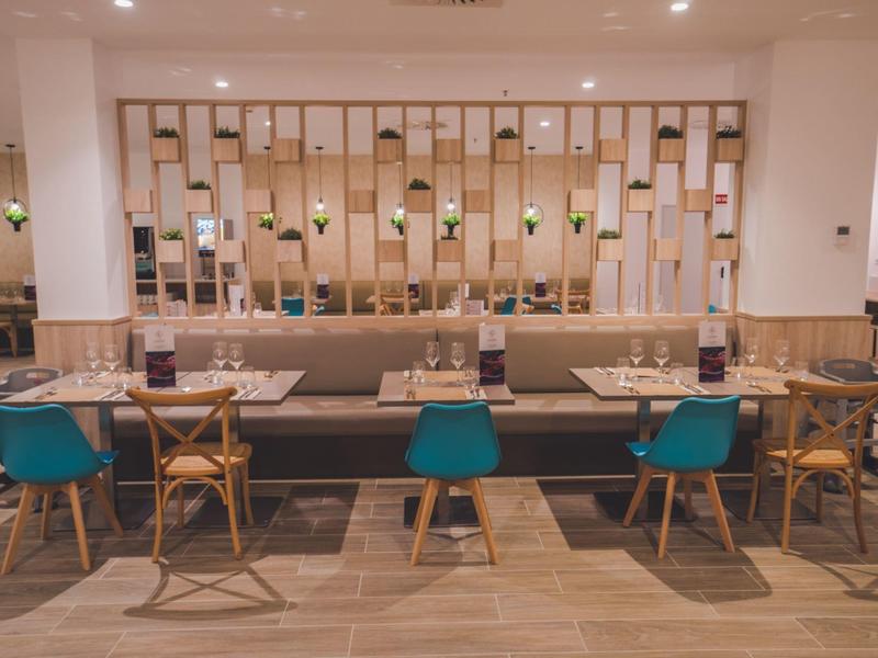 Nowoczesna jadalnia z drewnianymi stołami, kolorowymi krzesłami i dekoracyjną ścianą w tle.