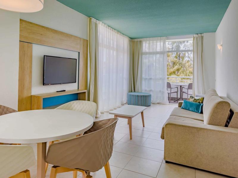 Jasny pokój hotelowy z częścią wypoczynkową, stołem jadalnym, telewizorem i balkonem z widokiem na zieleń.