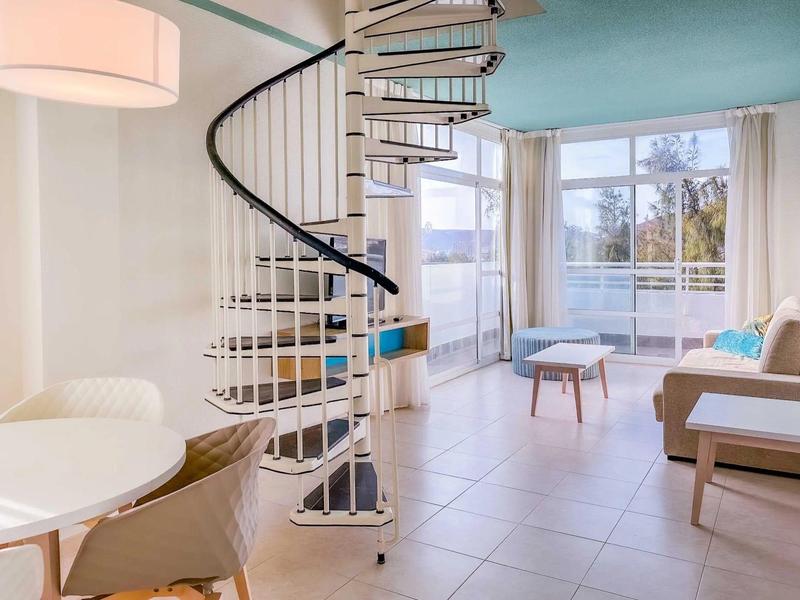 Jasny pokój dzienny ze spiralnymi schodami, dużymi oknami i niebieską sofą.