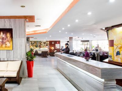 Bright hotel lobby with modern reception and seating area, decorated with artwork and plants.