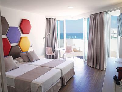 Bright hotel room with two beds, colorful wall decorations, and balcony with sea view.