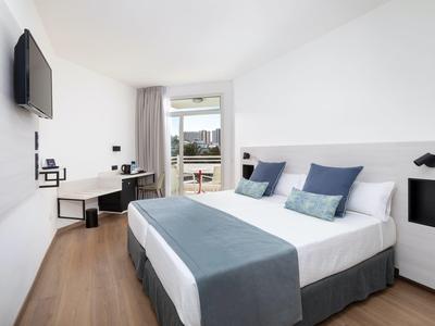 Bright hotel room with double bed, desk, and balcony overlooking the city.