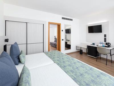 Modern hotel room with two single beds, desk, TV, and view into the adjoining room.