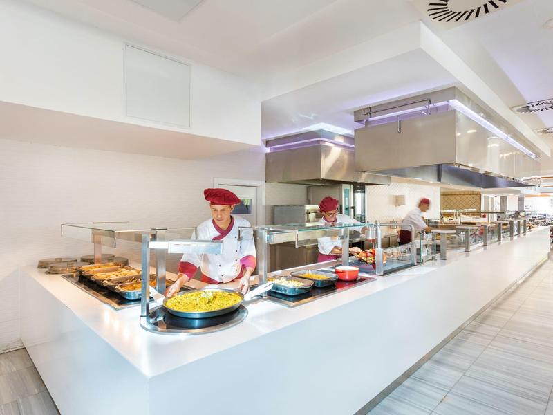 Bright cafeteria with long buffet and staff member in red uniform serving food.