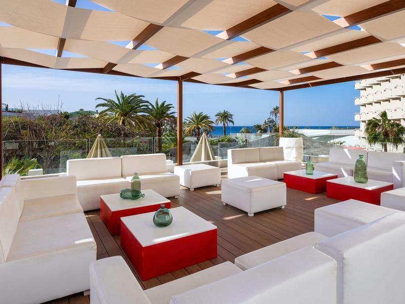Modern outdoor lounge with white sofas and red coffee tables, overlooking palm trees and the sea.
