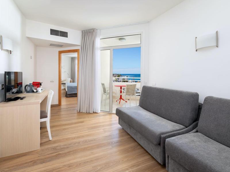 Bright hotel room with two gray sofas, TV, and balcony with sea view.