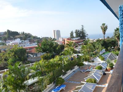 Balkon mit blauer Wand, Liegestühlen und Blick auf grüne Bäume, Gebäude und das Meer im Hintergrund.