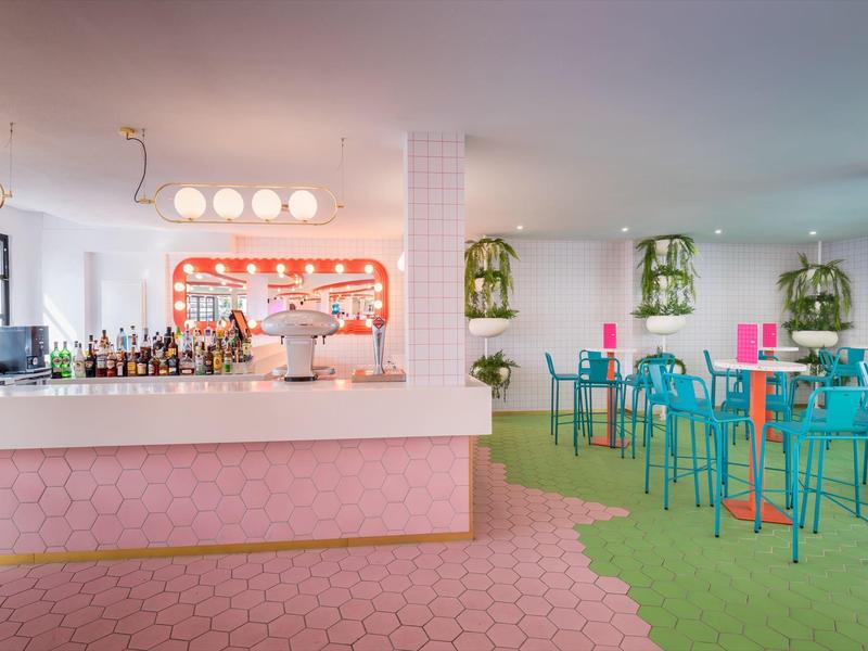 Nowoczesny bar z białym kontuarem, różowo-zieloną podłogą i niebieskimi krzesłami w jasnym pomieszczeniu.