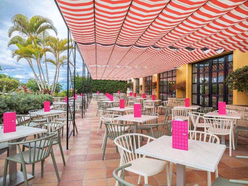 Zewnętrzny taras hotelowy z białymi stołami, szarymi krzesłami i czerwonymi markizami.