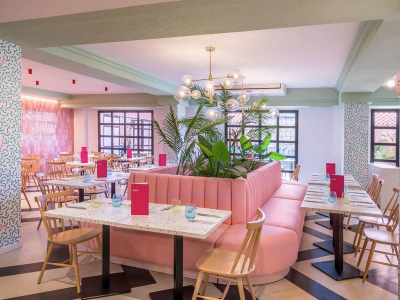 Jasna sala restauracyjna z różową kanapą, drewnianymi stołami i jasnymi krzesłami.