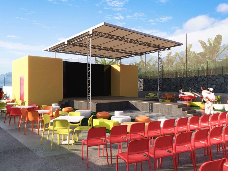 Zewnętrzny taras z kolorowymi krzesłami i zadaszoną sceną, widok na morze i góry.