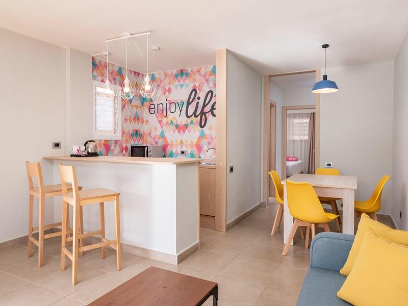 Jasny salon z żółtym kącikiem jadalnym i białym barem, kolorowa ściana z napisem "enjoy life".