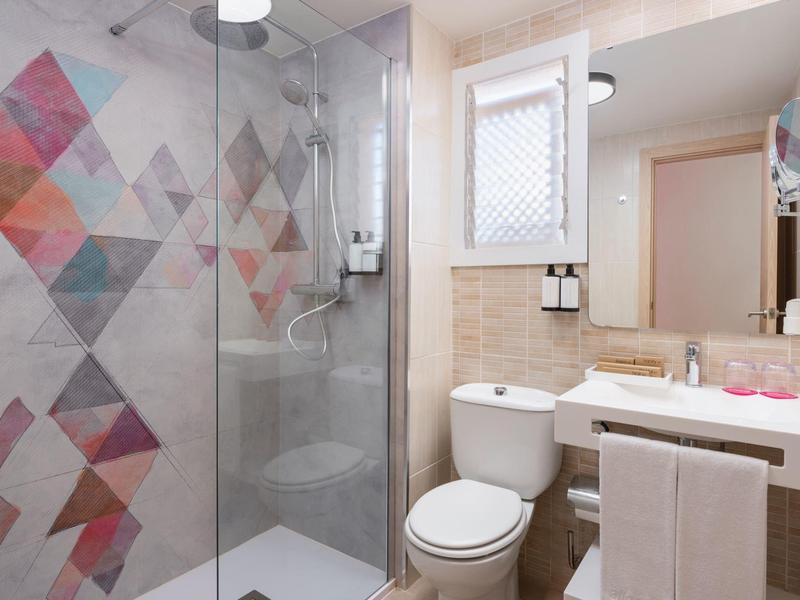 Łazienka z prysznicem z przeszkloną ścianą, kolorowymi płytkami, toaletą i umywalką z lustrem.