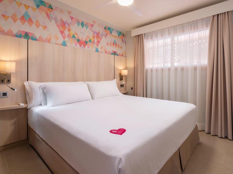 Nowoczesny pokój hotelowy z dużym łóżkiem, jasnymi ścianami i kolorową dekoracją.