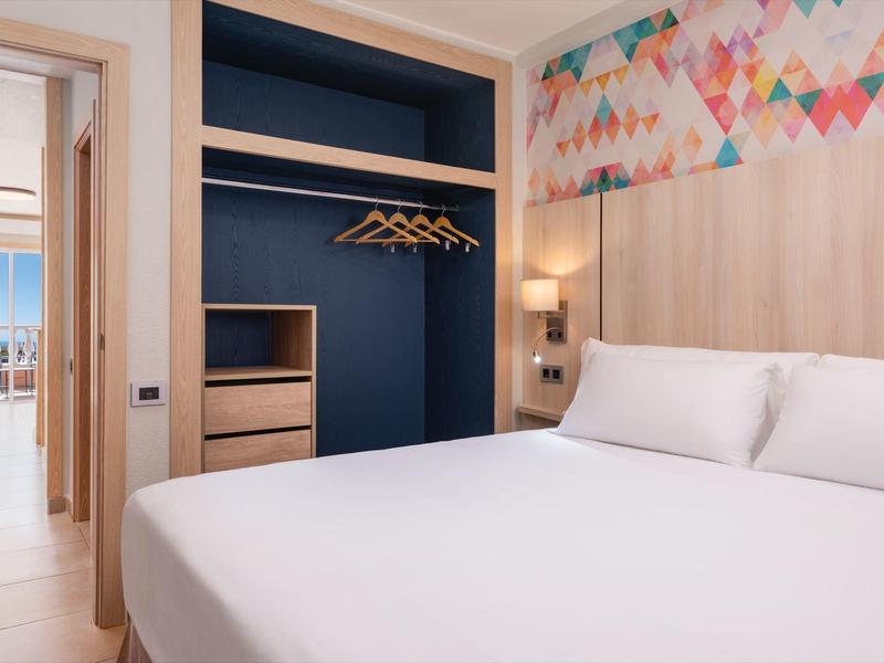 Nowoczesny pokój hotelowy z dużym łóżkiem, otwartą szafą i widokiem na morze.