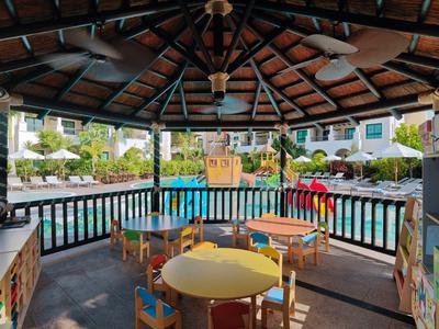 Overdekt terras met tafels en stoelen met uitzicht op een zwembad omgeven door groen.