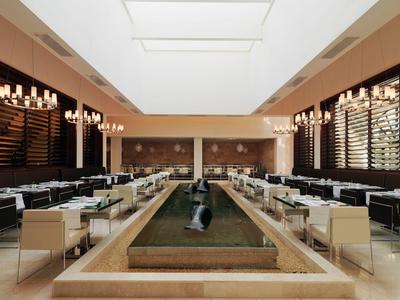 Moderne hotel eetzaal met symmetrische zitplaatsen, kroonluchters en centrale waterpartij onder dakramen.