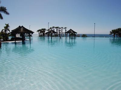 Infinity pool met houten bungalows boven het water en palmbomen bij een heldere hemel.