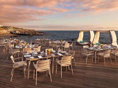 Buitenhouten terrasrestaurant met tafels en stoelen aan zee bij zonsondergang, zeilboten in het water.
