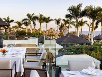 Buitenrestaurant met witte tafelkleden en palmbomen op de achtergrond bij zonsondergang.