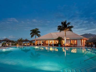 Groot buitenzwembad met helder blauw water en verlichte paviljoen onder schemerachtige lucht met palmbomen.