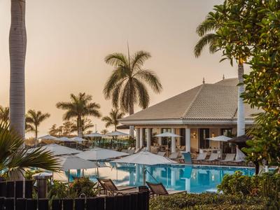 Ein Hotelpool mit blauen Liegestühlen, weißen Sonnenschirmen und Palmen bei Sonnenuntergang.