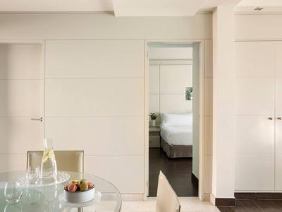 Moderne hotelkamer met glazen tafel, stoelen en slaapkamer zichtbaar door schuifdeur.