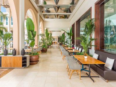 Buiten eetgedeelte van hotel met houten tafels, grijze stoelen en grote groene planten onder bogen.