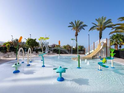 Buitenzwembad met waterstralen, glijbanen en palmbomen onder een heldere blauwe lucht.