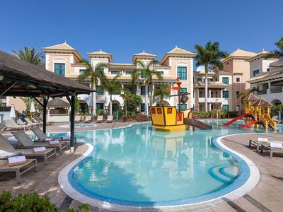 Hotelzwembad met speelse watervoorzieningen, ligstoelen en palmbomen onder een heldere blauwe lucht.