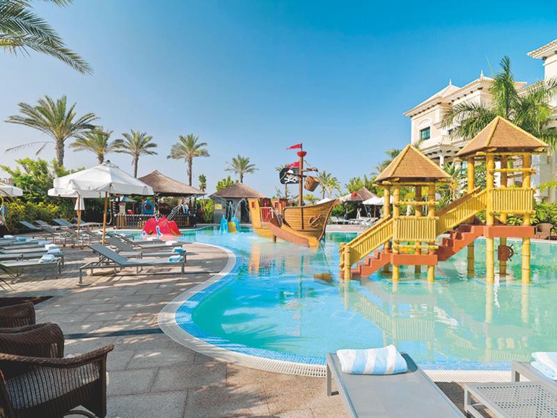 Resort zwembad met piratenschip-speeltuin en ligstoelen onder parasols op een zonnige dag.