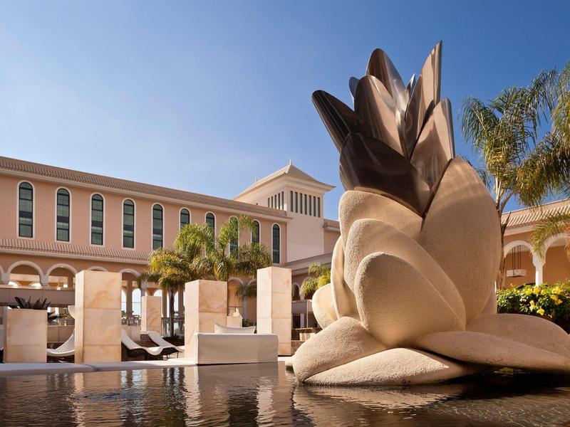 Hotelbinnenplaats met groot ananassculptuur, palmbomen, ligstoelen en heldere blauwe lucht.