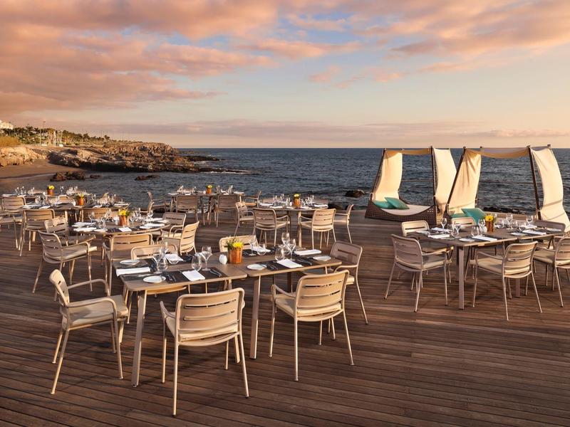 Buitenhouten terrasrestaurant met tafels en stoelen aan zee bij zonsondergang, zeilboten in het water.