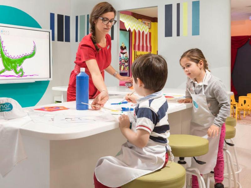 Kinderen en een volwassene aan een tafel bezig met een creatieve activiteit in een kleurrijke kamer.