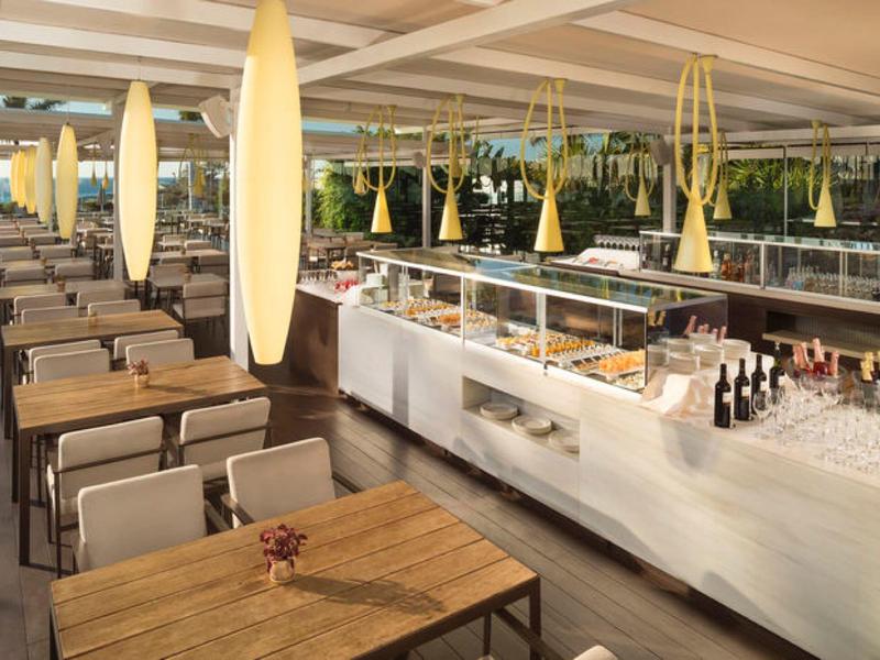 Buitenrestaurant met houten tafels, rieten stoelen en buffetbalie onder een overdekt terras.