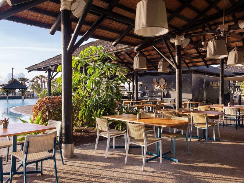 Buitenrestaurant met houten tafels en stoelen bij een zwembad en tropische planten.