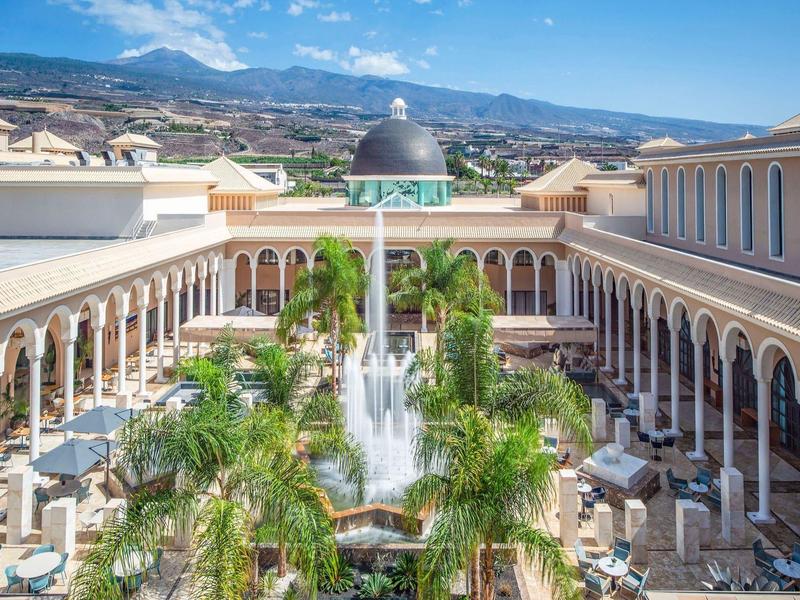 Ruime buitenhof met fontein, palmbomen en bogen onder een blauwe lucht.