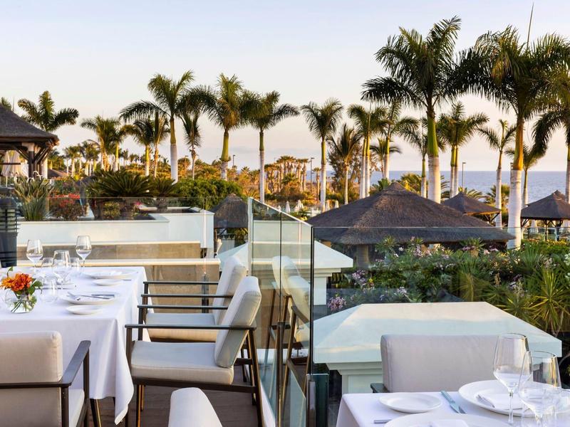 Buitenrestaurant met witte tafelkleden en palmbomen op de achtergrond bij zonsondergang.