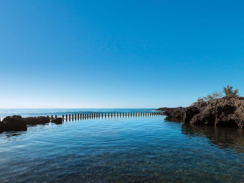 Heldere blauwe zee met rotsachtige kusten en een lange houten pier onder een heldere lucht.