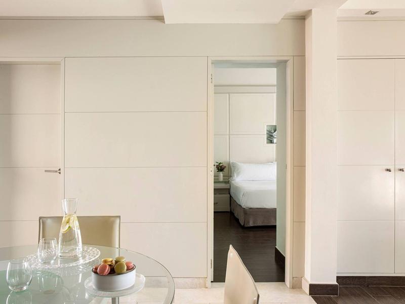 Moderne hotelkamer met glazen tafel, stoelen en slaapkamer zichtbaar door schuifdeur.