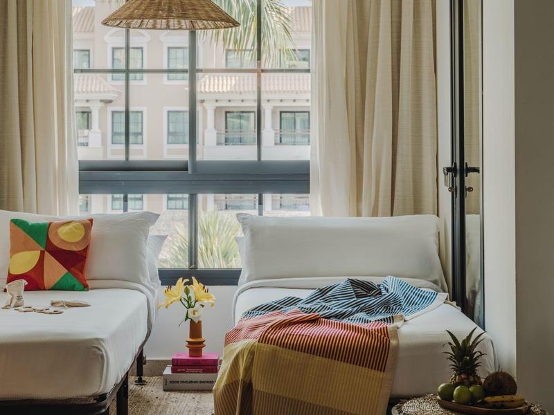Lichte hotelkamer met groot raam, gezellige banken, gestreept deken en decoratieve planten.