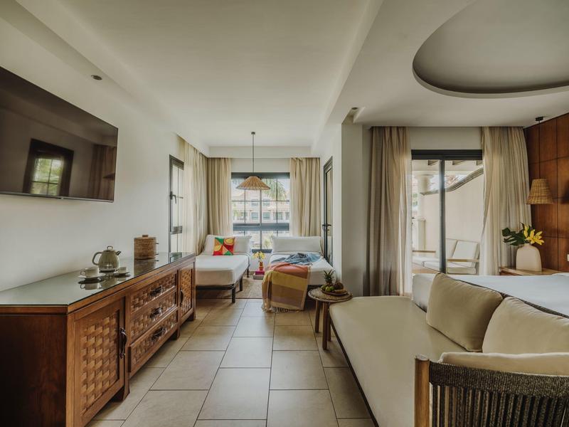 Lichte hotelkamer met bank, bed, tv en groot raam met gordijnen.